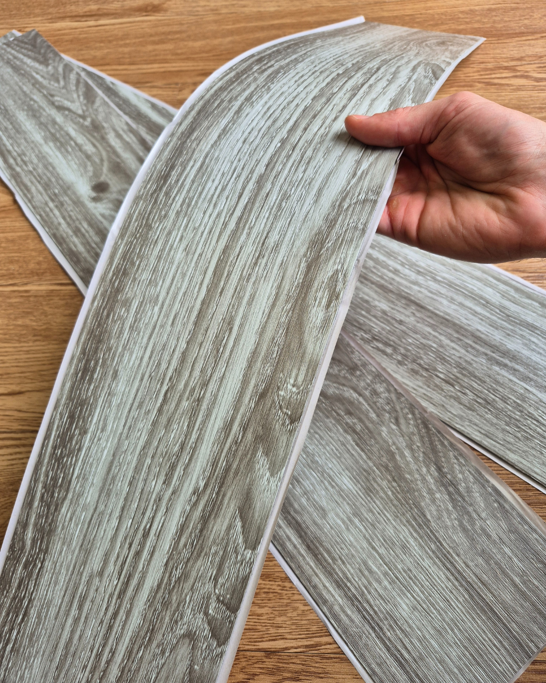 Wood-look vinyl flooring samples held by a hand on a wooden surface